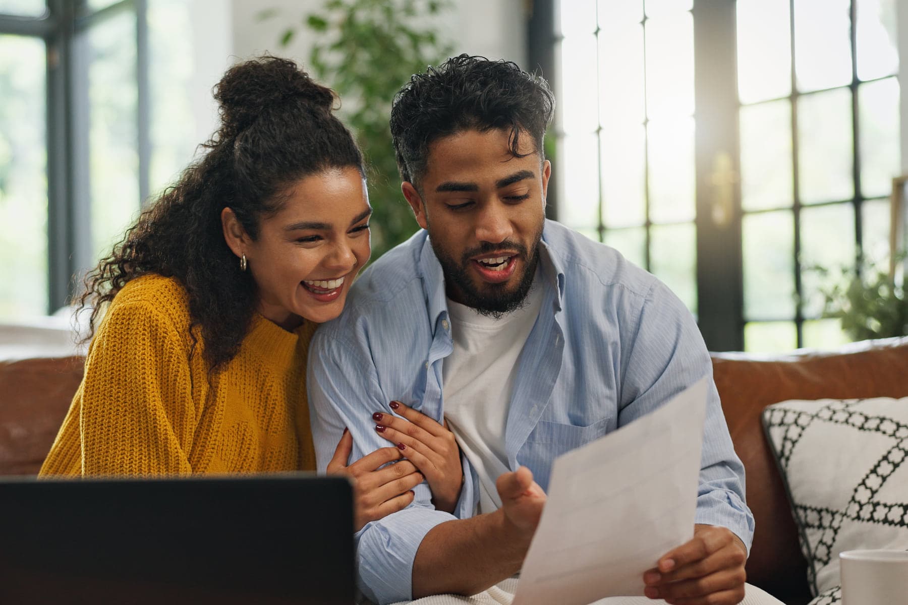 Two people sitting close together on a sofa at home, reading the same sheet of paper that one of them is holding up.
